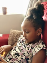 A child is sitting on a red sofa, engrossed in an activity, perhaps on a mobile device. The child is wearing a white t-shirt with a cartoon pattern featuring Minnie Mouse and pink floral accents. A brown leopard print pillow is visible in the background, and there is soft, natural lighting coming from a window nearby.