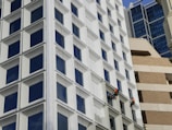 Two workers wearing safety gear and orange vests are suspended on the side of a modern building, painting the exterior. The building features a grid of large, square windows with white frames. The background is a mix of different architectural styles, including glass and brick structures.