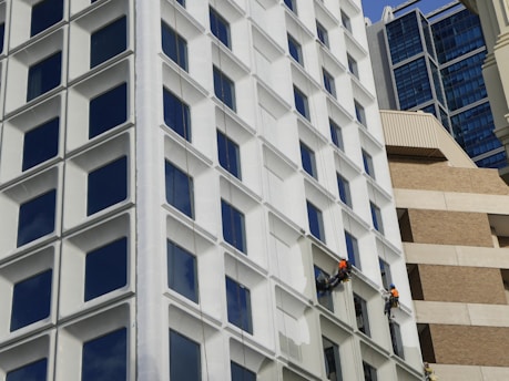 Two workers wearing safety gear and orange vests are suspended on the side of a modern building, painting the exterior. The building features a grid of large, square windows with white frames. The background is a mix of different architectural styles, including glass and brick structures.