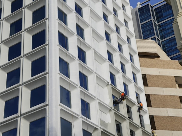 Two workers wearing safety gear and orange vests are suspended on the side of a modern building, painting the exterior. The building features a grid of large, square windows with white frames. The background is a mix of different architectural styles, including glass and brick structures.