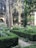 A beautifully maintained garden featuring trimmed hedges, tall trees, and a central fountain surrounded by lush greenery. People are visible in the background, partially obscured by the foliage. The garden is enclosed by a building with arched windows and decorative columns.