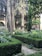 A beautifully maintained garden featuring trimmed hedges, tall trees, and a central fountain surrounded by lush greenery. People are visible in the background, partially obscured by the foliage. The garden is enclosed by a building with arched windows and decorative columns.