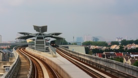 A modern train station with a sleek, futuristic design is visible, featuring multiple steel beams and a large canopy. The railway tracks curve gently towards the station. Surrounding the station are rows of houses with slanted rooftops, and in the background, several high-rise buildings are visible under a cloudy sky.