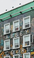 The facade of a building features the words 'Abbey Court' prominently displayed in large white letters. The exterior is covered in intricate graffiti and artwork depicting colorful flowers and abstract designs surrounding the windows. The upper part of the building is painted in green, and there is a 'Vacancies' sign to the right of the windows.