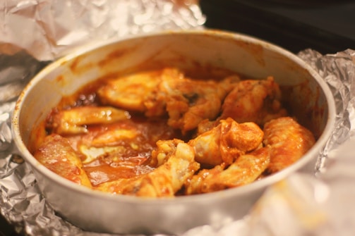 A dish containing several pieces of cooked chicken wings is placed in a circular metal baking pan. The pan is surrounded by crumpled aluminum foil, and the chicken pieces are coated in a rich, red-brown sauce.
