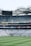 A large stadium with empty seating sections and a well-maintained grass field. The seats are mostly blue and are arranged in tiers, with a scoreboard or large screen positioned at the top. Advertising banners and signs are visible along the stands, and the stadium is covered by a partially translucent roof structure.