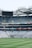 A large stadium with empty seating sections and a well-maintained grass field. The seats are mostly blue and are arranged in tiers, with a scoreboard or large screen positioned at the top. Advertising banners and signs are visible along the stands, and the stadium is covered by a partially translucent roof structure.