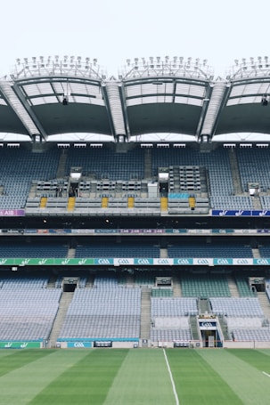 A large, empty stadium with multiple tiers of seating, featuring a well-maintained green field in the foreground. The stadium's roof is modern and arching, with lighting fixtures visible on the top. Advertising boards and signage are displayed along the tiers.