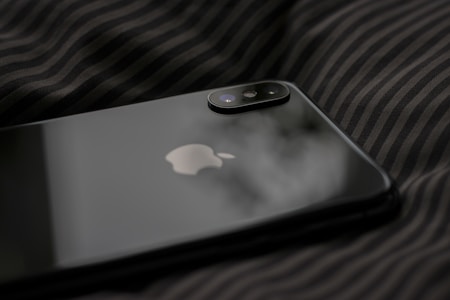 A close-up view of a sleek, black smartphone with Apple's logo on its back panel. The phone is resting on a dark, striped fabric surface. The dual camera setup on the phone is prominently visible.