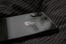 A close-up view of a sleek, black smartphone with Apple's logo on its back panel. The phone is resting on a dark, striped fabric surface. The dual camera setup on the phone is prominently visible.