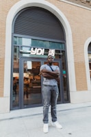 A man stands confidently with folded arms in front of a modern storefront with large glass doors. The building features a brick facade and an arched window above. The interior of the store is softly lit, showcasing various items. The man is casually dressed in a gray T-shirt, distressed jeans, and white sneakers.