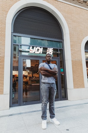 A man stands confidently with folded arms in front of a modern storefront with large glass doors. The building features a brick facade and an arched window above. The interior of the store is softly lit, showcasing various items. The man is casually dressed in a gray T-shirt, distressed jeans, and white sneakers.