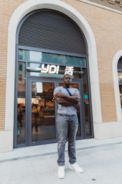A man stands confidently with folded arms in front of a modern storefront with large glass doors. The building features a brick facade and an arched window above. The interior of the store is softly lit, showcasing various items. The man is casually dressed in a gray T-shirt, distressed jeans, and white sneakers.