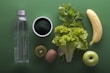 A neatly arranged assortment of healthy foods sits on a green background, including a clear water bottle, a bowl of dark green powder, leafy celery and lettuce, a banana, sliced kiwi, and a green apple.