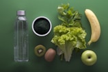 A neatly arranged assortment of healthy foods sits on a green background, including a clear water bottle, a bowl of dark green powder, leafy celery and lettuce, a banana, sliced kiwi, and a green apple.