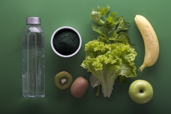 A neatly arranged assortment of healthy foods sits on a green background, including a clear water bottle, a bowl of dark green powder, leafy celery and lettuce, a banana, sliced kiwi, and a green apple.
