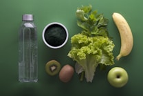 A neatly arranged assortment of healthy foods sits on a green background, including a clear water bottle, a bowl of dark green powder, leafy celery and lettuce, a banana, sliced kiwi, and a green apple.