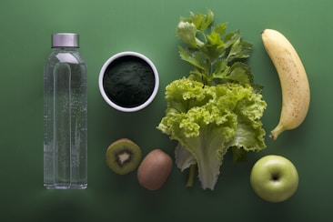 A neatly arranged assortment of healthy foods sits on a green background, including a clear water bottle, a bowl of dark green powder, leafy celery and lettuce, a banana, sliced kiwi, and a green apple.