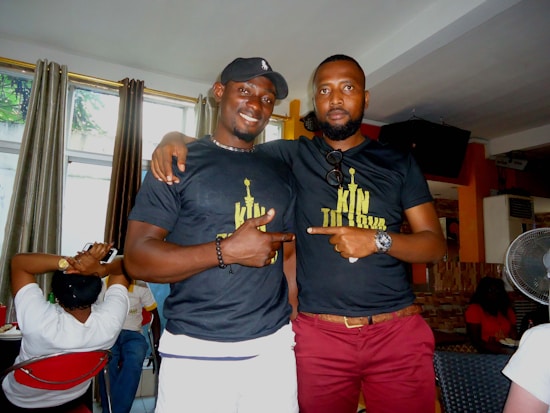Two men wearing matching black t-shirts stand together indoors with one arm around each other. They both point to the design on their shirts, and the room has curtains, a television, and several other people seated in the background.