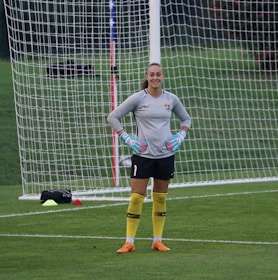 A soccer goalkeeper stands on a field in front of a goal net, wearing bright yellow socks, orange cleats, and a grey long-sleeve jersey. The person has blue and pink goalkeeper gloves and appears to be smiling.