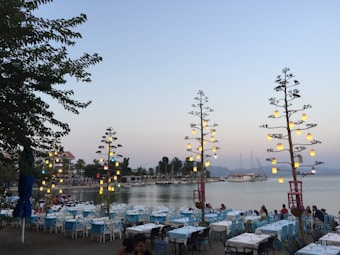 A waterfront dining area features rows of tables with white tablecloths and aqua blue chairs set near the shore. Decorative pole lights with hanging lanterns illuminate the scene, adding a warm glow. In the background, several boats are moored, with a backdrop of calm water and distant hills. Trees provide additional greenery, while people can be seen enjoying the outdoor setting.