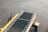 A conveyor belt system with yellow side rails is positioned on a concrete surface, likely outdoors. The apparatus has a black rubber belt with visible metallic rollers along the edges. Adjacent to the conveyor is a small step with a metal mesh texture.