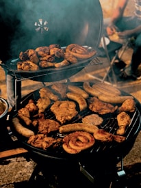 Various types of meat, including sausages and chicken, are being grilled on a barbecue. Smoke is rising from the grill, creating an inviting and rustic atmosphere. In the background, partially visible people are seated on chairs.