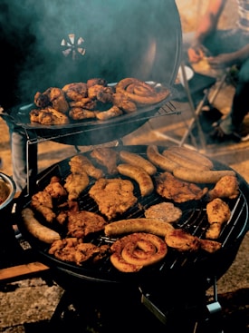 Various types of meat, including sausages and chicken, are being grilled on a barbecue. Smoke is rising from the grill, creating an inviting and rustic atmosphere. In the background, partially visible people are seated on chairs.