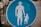 A circular blue sign with white silhouettes of an adult holding hands with a child is prominently displayed. The sign is placed on a sidewalk with some greenery on the left side. The background shows part of an urban environment with pavement and blurred elements in the distance.