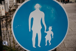 A circular blue sign with white silhouettes of an adult holding hands with a child is prominently displayed. The sign is placed on a sidewalk with some greenery on the left side. The background shows part of an urban environment with pavement and blurred elements in the distance.