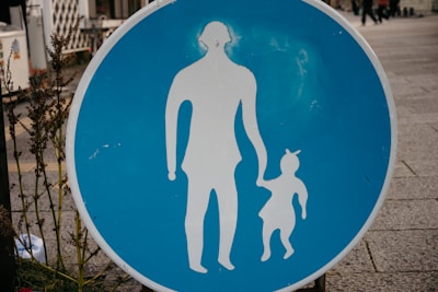 A circular blue sign with white silhouettes of an adult holding hands with a child is prominently displayed. The sign is placed on a sidewalk with some greenery on the left side. The background shows part of an urban environment with pavement and blurred elements in the distance.