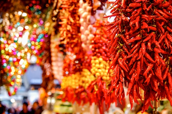 Bundles of red chili peppers are hanging next to clusters of garlic and other produce in a vibrant market setting. The background features blurred colorful lights and other hanging decorations, creating a festive atmosphere.