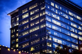 A modern glass office building illuminated from within, set against a twilight sky. Light reflects off the many windows, creating a grid-like pattern. Some architectural details are visible, including corner elements and external lighting.