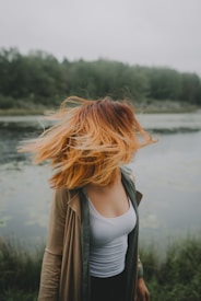 A person stands near a tranquil body of water, with their hair catching a breeze and obscuring their face. Surrounded by lush greenery, the scene conveys a sense of freedom and connection with nature.