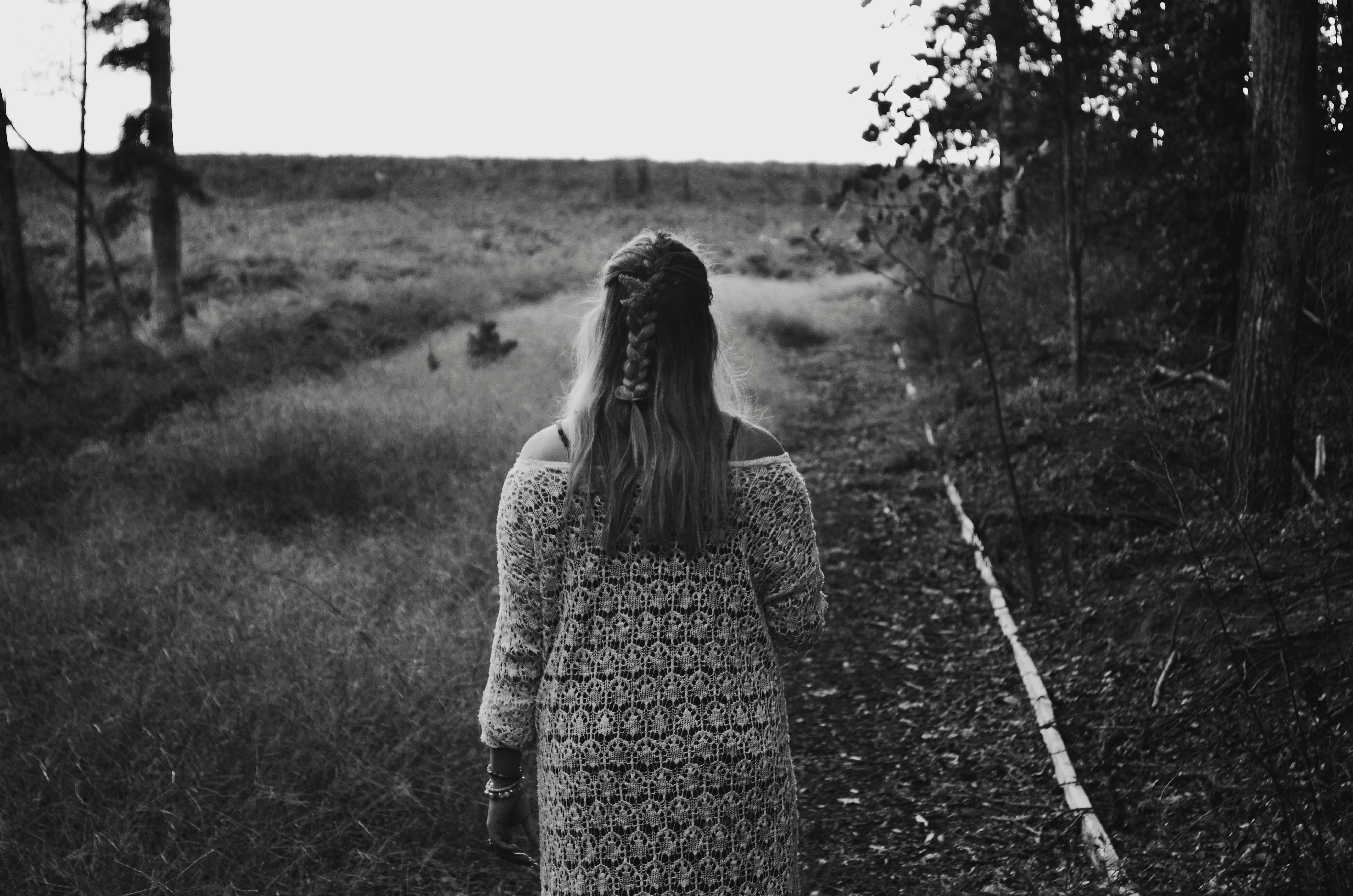 grayscale photo of woman wearing dress walking across grass field between trees
