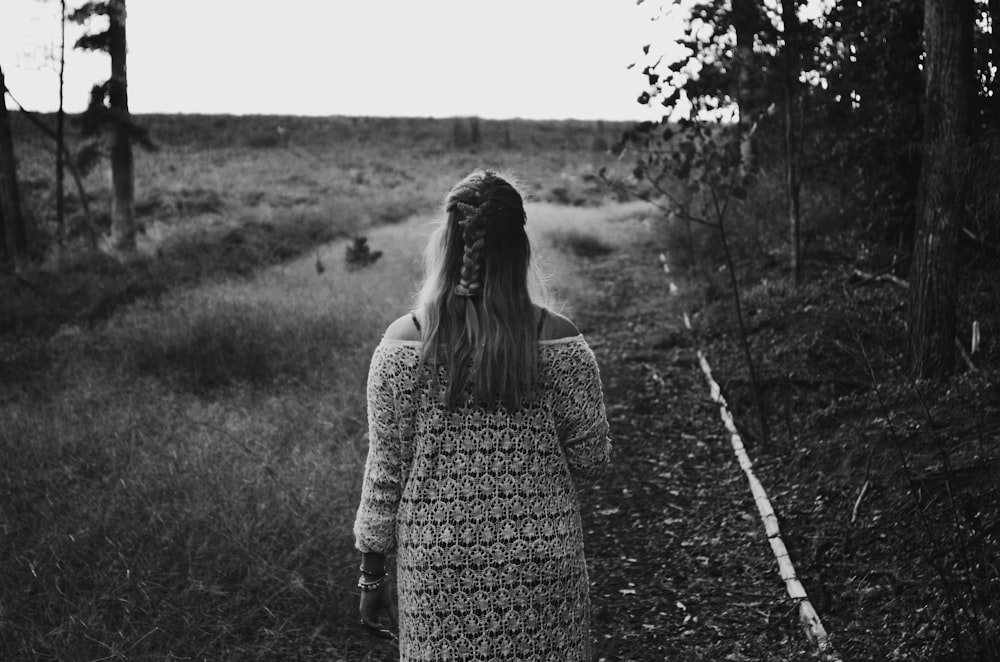 grayscale photo of woman wearing dress walking across grass field between trees