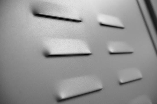 A close-up view of a metal surface with rectangular vents. The vents are evenly spaced and create a subtle pattern on the surface. The lighting highlights the texture and creates soft shadows.