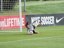 A goalkeeper is actively diving toward a soccer ball, making a save near the goalpost on a grassy field. The advertising boards in the background display branding related to soccer.
