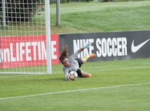 A goalkeeper is actively diving toward a soccer ball, making a save near the goalpost on a grassy field. The advertising boards in the background display branding related to soccer.
