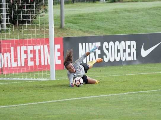 A goalkeeper is actively diving toward a soccer ball, making a save near the goalpost on a grassy field. The advertising boards in the background display branding related to soccer.