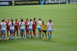 A group of female soccer players are standing on the field. One team is wearing red and black jerseys, while the other team is wearing white jerseys with light blue shorts. The players are interacting with each other, possibly before or after a match. The field is lush green, and there are banners and advertisements on the sidelines.