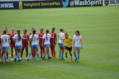 A group of female soccer players are standing on the field. One team is wearing red and black jerseys, while the other team is wearing white jerseys with light blue shorts. The players are interacting with each other, possibly before or after a match. The field is lush green, and there are banners and advertisements on the sidelines.