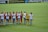 A group of female soccer players are standing on the field. One team is wearing red and black jerseys, while the other team is wearing white jerseys with light blue shorts. The players are interacting with each other, possibly before or after a match. The field is lush green, and there are banners and advertisements on the sidelines.