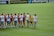 A group of female soccer players are standing on the field. One team is wearing red and black jerseys, while the other team is wearing white jerseys with light blue shorts. The players are interacting with each other, possibly before or after a match. The field is lush green, and there are banners and advertisements on the sidelines.