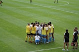 A group of female soccer players, wearing yellow and white jerseys, stands in a tight huddle on a lush green soccer field. The players are focused and engaged, suggesting they are discussing strategies or motivating each other. Nearby, two coaches in black sportswear observe and provide guidance.