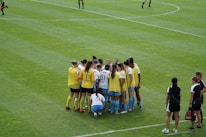 A group of female soccer players, wearing yellow and white jerseys, stands in a tight huddle on a lush green soccer field. The players are focused and engaged, suggesting they are discussing strategies or motivating each other. Nearby, two coaches in black sportswear observe and provide guidance.