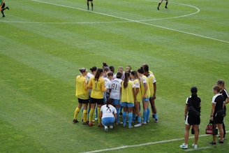 A group of female soccer players, wearing yellow and white jerseys, stands in a tight huddle on a lush green soccer field. The players are focused and engaged, suggesting they are discussing strategies or motivating each other. Nearby, two coaches in black sportswear observe and provide guidance.
