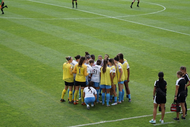 A group of female soccer players, wearing yellow and white jerseys, stands in a tight huddle on a lush green soccer field. The players are focused and engaged, suggesting they are discussing strategies or motivating each other. Nearby, two coaches in black sportswear observe and provide guidance.