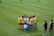 A group of female soccer players, wearing yellow and white jerseys, stands in a tight huddle on a lush green soccer field. The players are focused and engaged, suggesting they are discussing strategies or motivating each other. Nearby, two coaches in black sportswear observe and provide guidance.