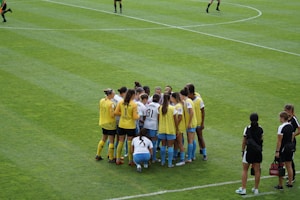A group of female soccer players, wearing yellow and white jerseys, stands in a tight huddle on a lush green soccer field. The players are focused and engaged, suggesting they are discussing strategies or motivating each other. Nearby, two coaches in black sportswear observe and provide guidance.